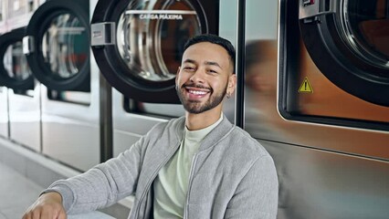 Wall Mural - Young hispanic man smiling confident sitting on the floor at laundry facility