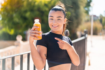 Sticker - Young pretty sport woman holding an orange juice at outdoors and pointing it