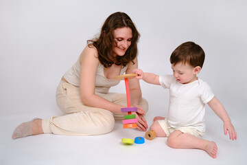 Wall Mural - Happy baby with mother play educational toys on studio white background. Portrait of a smiling child with mom and playing while sitting on the floor. Kid about two years old (one year nine months)