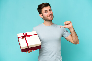 Wall Mural - Young caucasian man holding a gift isolated on blue background proud and self-satisfied