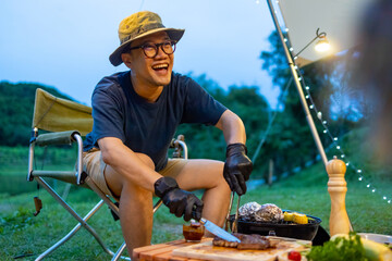 Wall Mural - Asian man cooking meal grilled bbq steak for dinner party at camp at sunset. Group of People enjoy outdoor adventure lifestyle travel nature hiking and camping together on summer holiday vacation.