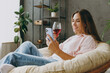 © ViDi Studio - Side view young woman wear casual clothes sits in armchair hold wineglass drink wine use mobile cell phone stay home hotel flat rest relax spend free spare time in living room indoor Lounge concept.