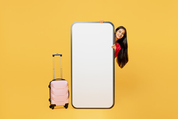 Wall Mural - Traveler Indian woman hold suitcase big huge blank screen mobile cell phone isolated on plain yellow background. Tourist travel abroad in free spare time rest getaway. Air flight trip journey concept.