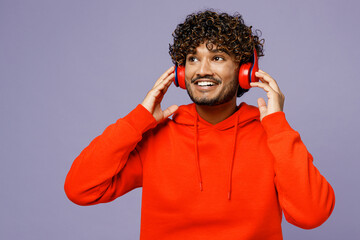 Poster - Young fun Indian man wears red orange hoody casual clothes listen to music in headphones look aside on area isolated on plain pastel light purple color background studio portrait. Lifestyle concept.