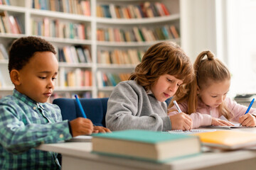 Poster - Smart diverse schoolchildren sitting at table in classroom at primary school, writing or drawing in notebooks