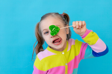 portrait of a cute little girl with lollipops in a striped jacket covered one eye with a large candy on a stick. The concept of sweets and confectionery. Photo studio, blue background, place for text