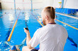 A young swimming coach with a stopwatch in his hand watches the training session near the sports pool.
