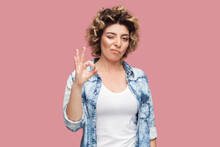 Portrait Of Playful Woman With Curly Hairstyle Wearing Blue Shirt Standing Showing Okay Gesture, Winking To Camera, Saying Great Job. Indoor Studio Shot Isolated On Pink Background.
