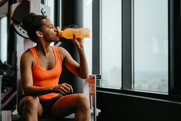 Young fit woman wearing sports clothes sitting in the fitness gym near window and drinking water from a bottle after doing an exercise.