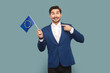 © khosrork - Portrait of excited amazed man with mustache standing pointing at Europe flag, looking at camera, wearing official style suit. Indoor studio shot isolated on light blue background.