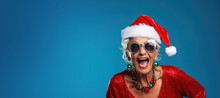Happy Laughing Mature Woman Wearing A Santa Hat For Christmas On A Blue Background With Space For Copy