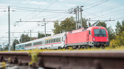 Wall Mural - Passenger train with modern red locomotive and four coaches is rushing through the station of Ljubljana Vizmarje on a summer day. Low profile photo.