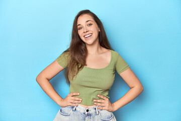 Young Caucasian woman in a green top on a blue backdrop confident keeping hands on hips.