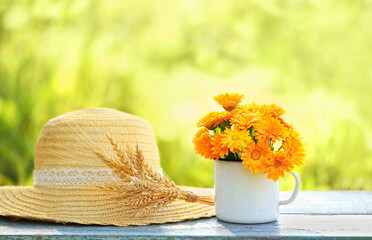 Poster - summer background. calendula flowers in enamel cup and rustic braided hat with wheat ears on table in garden, natural abstract background. summer seasonal composition with flowers. template for design