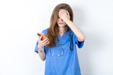 Young Caucasian Doctor Woman Wearing Medical Uniform  Looking At Smart Phone Feeling Sad Holding Hand On Face.