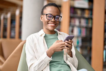 Happy African Black teen girl college generation z student wearing eyeglasses with smartphone looking at camera using mobile cell phone modern tech device sitting in university campus. Portrait.