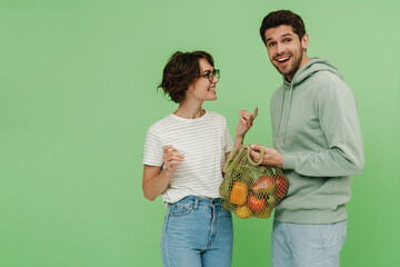 Smiling man and woman holding mesh shopping bag with fruits isolated over green wall