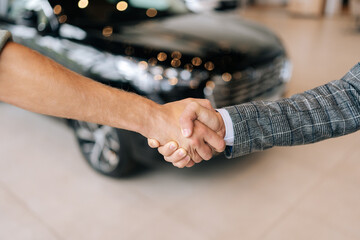 Wall Mural - Closeup cropped shot of successful purchase of vehicle, unrecognizable satisfied buyers shake hands with male seller after buying new car in auto salon. Concept of choosing buying new car at showroom.