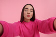 Young woman taking selfie and blowing kiss on pink background