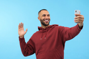 Wall Mural - Smiling young man taking selfie with smartphone on light blue background