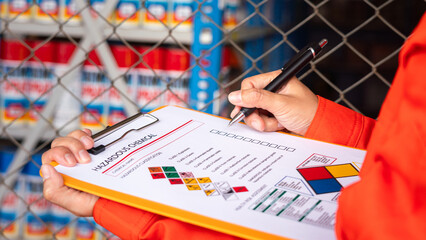 A worker is checking on the hazardous chemical material information form with background of chemical storage area at the factory place. Industrial safety working action. Selective focus.