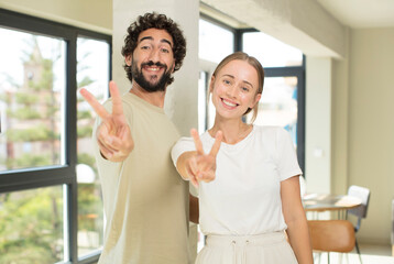 Canvas Print - young adult couple smiling and looking happy, carefree and positive, gesturing victory or peace with one hand