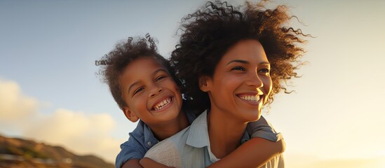 happy african american mother carrying her son on her back at the beach under a blue sky during suns