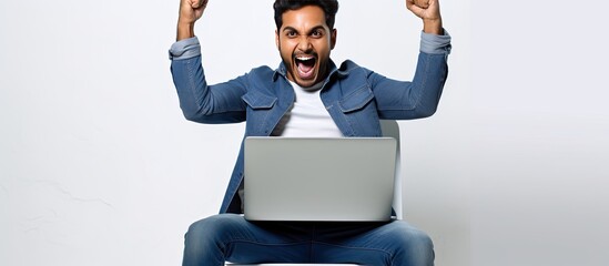 Enthusiastic Indian man celebrating success on laptop sitting on chair against white wall background looking at computer screen and shaking fists happily