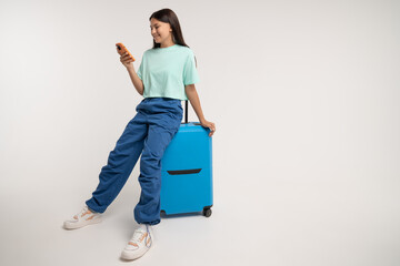 Smiling teenage girl sits on travel blue suitcase and using smartphone. Happy teen girl tourist inspired by upcoming trip chooses route on phone isolated on gray studio background with copy space