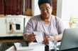 Portrait of puzzled upset serious overweight african american grandma in glasses reading paycheck or utility bill attentively sitting in her kitchen in front of laptop and smartphone