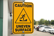 Caution Uneven Surface Sign With Caption And Illustration Picture Of Person Walking On An Uneven Surface Surrounded By A Triangle, Building Wall And Parking Lot With Parked Cars And Sky In Background