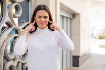 Wall Mural - pretty young adult woman looking angry, stressed and annoyed, covering both ears to a deafening noise, sound or loud music