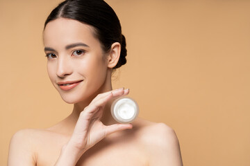 Canvas Print - Happy asian woman with naked shoulders and natural makeup holding cosmetic cream isolated on beige