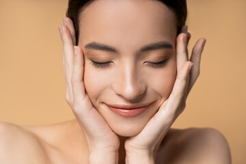 Wall Mural - Close up view of asian woman with closed eyes and naked shoulders touching face isolated on beige