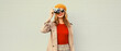 Fashionable autumn color style outfit, stylish young woman photographer with film camera wearing orange french beret hat, jacket and round sunglasses on gray background