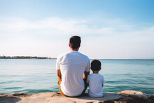 Young Father With His Son Wearing White T-shirt Sitting On Shore Of Sea Or Ocean. Rear View. Mock Up Template For T-shirt Design Print