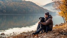 A Young Guy With His Black Labrador Dog Sits Hugging On The Shore Of A Mountain Lake On An Autumn Evening.