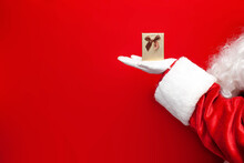 Small Gift Box In The Hands Of Santa Claus On Red Background, Santa Gives Festive Gift For The New Year