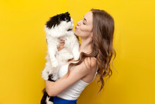 Young Cute Girl Kisses Black And White Cat On Yellow Isolated Background And Smiles, Woman With Pet