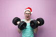 © Богдан Маліцький - young nerd guy in glasses and santa hat trains and lifts heavy dumbbells on pink isolated background