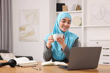 Wall Mural - Muslim woman in hijab with cup of coffee using laptop at wooden table in room