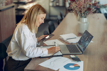 Wall Mural - Smiling business woman working on laptop, making notes in cozy coworkign space interior