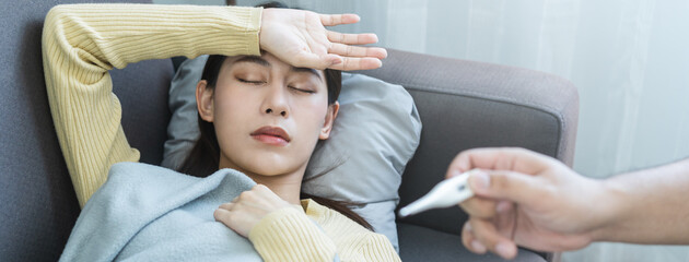 Wall Mural - Sick, influenza asian young woman, girl headache have a fever, flu and check thermometer measure body temperature, feel illness sitting on sofa bed at home. Health care person on virus, covid-19.