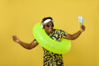Cheerful African-American man dancing while listening music on headphones with tickets in hand before traveling. perky dark-skinned guy in sunglasses and with inflatable circle goes on trip by plane