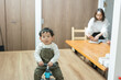 春の昼に部屋で三輪車にまたがって正面を見る日本人の子と母親
