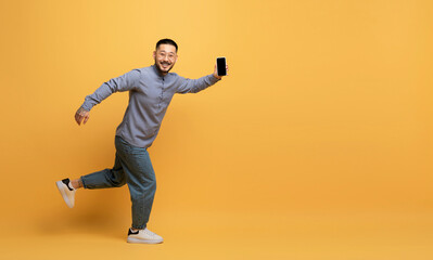 Poster - Cheerful young asian guy with blank smartphone running on yellow studio background