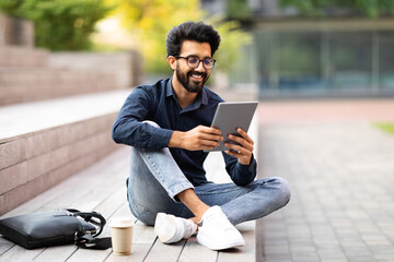 Wall Mural - Positive indian guy resting at park after work, using tablet