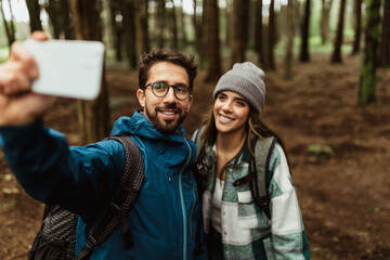 Wall Mural - Cheerful young caucasian couple in jackets in forest, make selfie on smartphone, enjoy vacation