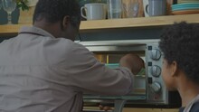 Senior African American Grandfather Putting Silicone Mold With Cake Batter Into Mini Oven, Then Smiling And Giving Fist Bump To Little Grandson While Baking Together In Kitchen