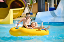 Boy And Girl Have Fun On Water Slide In Outdoor Aquapark. Little Children Floating On Yellow Inflatable Raft. Summer Water Play For The Kids.
